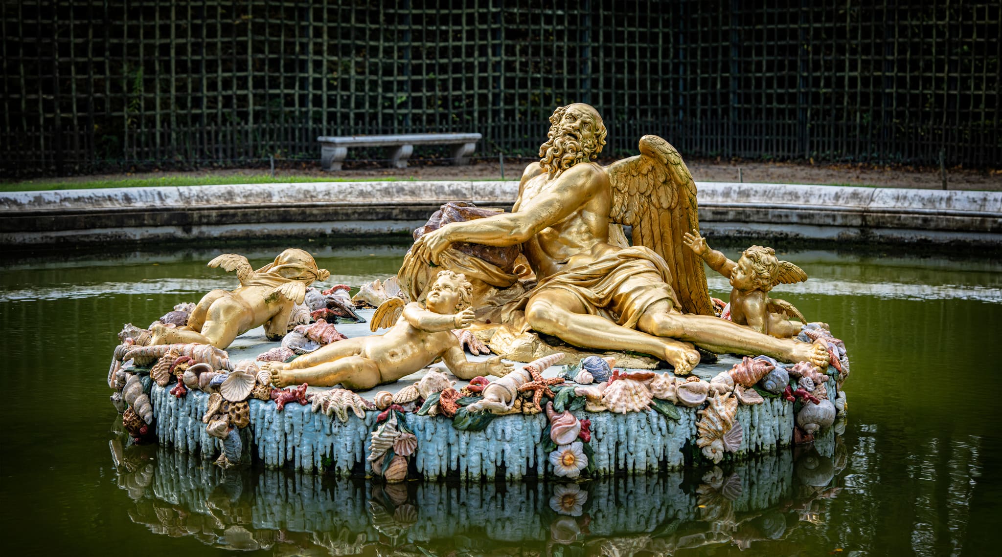 Gold statue of a Saturn in a fountain with water and green fenced background.